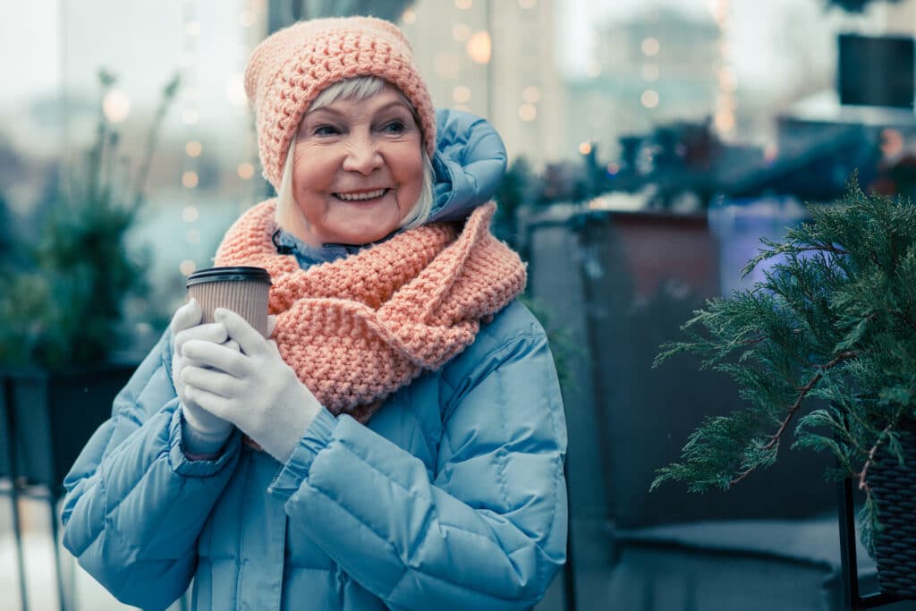 Cheerful aged woman spending time outdoors in a cold winter day and smiling while holding a cup of coffee